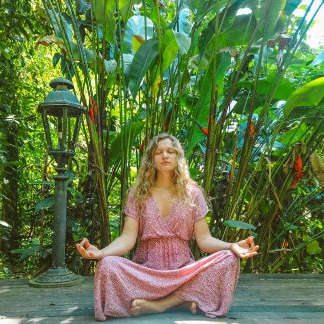 Woman meditating outdoors during a luxury wellness retreat in Rishikesh surrounded by tropical greenery, promoting yoga, mindfulness, relaxation, and holistic healing.